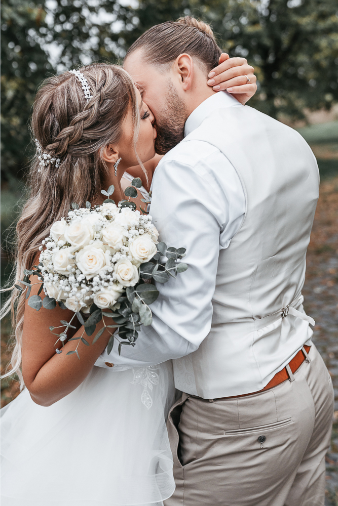 Brautpaar küsst sich nach der Hochzeit in Wolfsburg mit modernem Brautstrauß aus weißen Rosen und Eukalyptus.