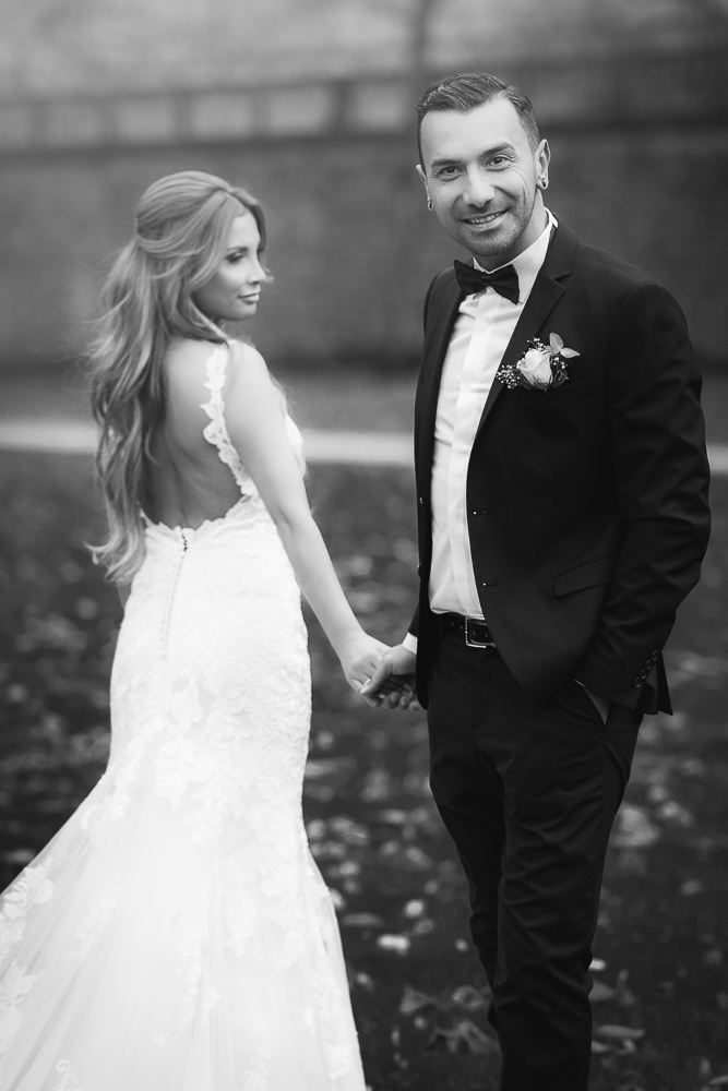Braut im Fit & Flare Brautkleid mit dem Rücken zum Betrachter, Händchenhaltend mit dem Bräutigam, der den Betrachter anschaut, Hochzeit in Wolfsburg.